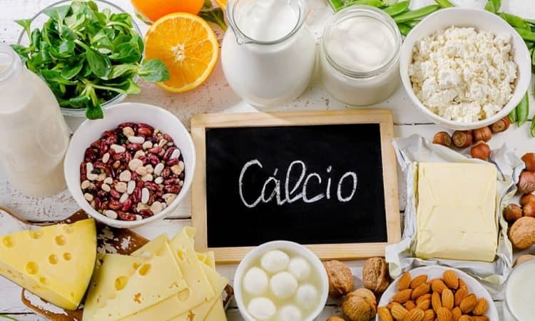 Alimentos ricos em cálcio, como queijos, leite, amêndoas, entre vários outros, espalhados em uma mesa. No centro da imagem, uma pequena lousa tem a palavra "cálcio" escrita.