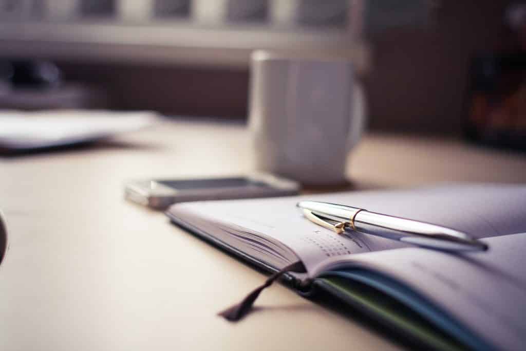 Mesa com caderno e caneta ao lado de uma xícara.