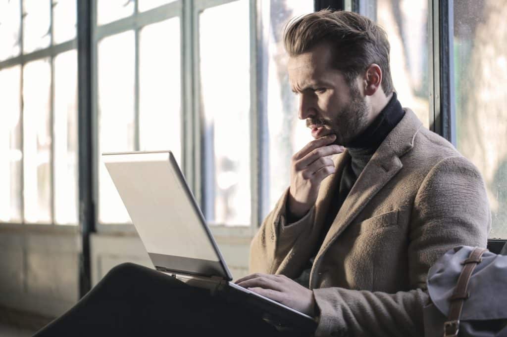 Homem com a mão direita no queixo e um notebook no colo, com a expressão pensativa enquanto olha para a tela.