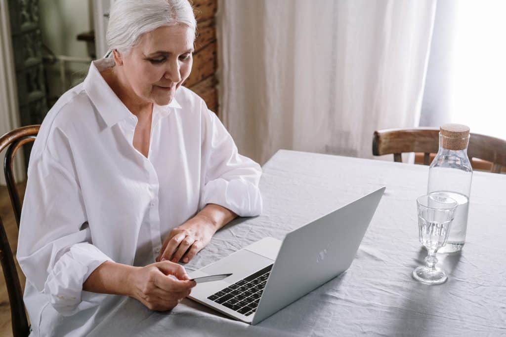 Mulher idosa sentada em uma mesa, usando o computador, que está ao lado de uma garrafa d'água.