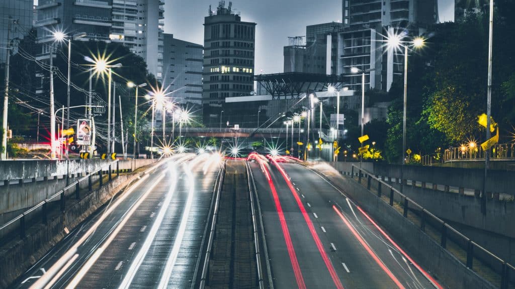 Foto em time lapse de uma rua movimentada ao começo da noite, com as luzes dos carros e dos postes de luz acesas.
