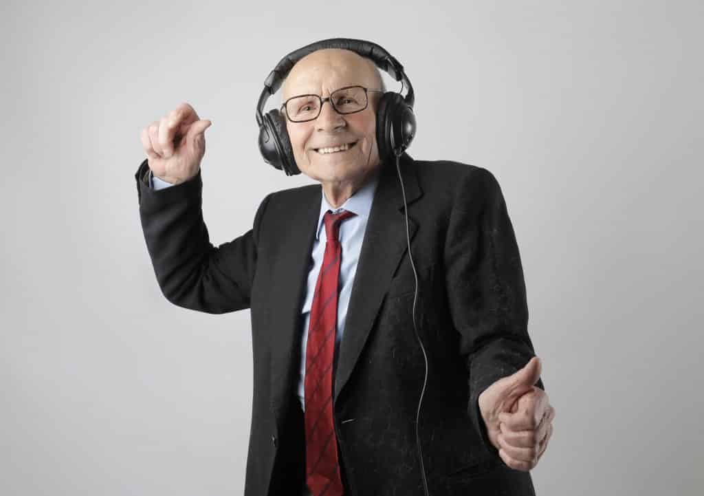 Homem idoso com roupas sociais dançando e sorrindo, com fones de ouvido.