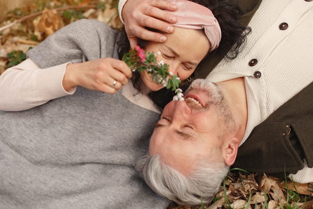 Casal de homem e mulher idosos deitado em um chão repleto de folhas, abraçados, sorrindo um para o outro.