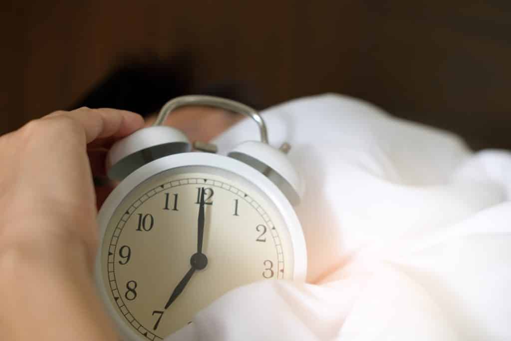 Pessoa segurando um relógio alarme na cama, sobre cobertores, que marca o horário das sete da manhã.
