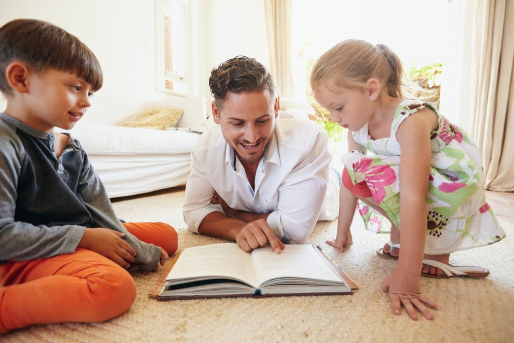 Family reading a book on the floor in the living room. Father reading stories to his son and daughter at home. Young man little boy and little girl reading story book.