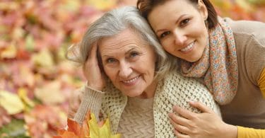 Mãe idosa e filha abraçadas, sorrindo, com folhas de outono ao fundo.