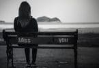 Mulher sentada em um banco em frente ao mar. No banco estão escritas as palavras "Miss" onde ela está sentada e "You" na ponta oposta do banco. "Miss You" é traduzido como "Sinto sua falta" em português.