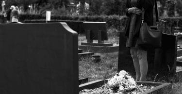 Mulher em pé em frente a um túmulo em um cemitério, demonstrando luto e tristeza.. Sua mão está cobrindo o rosto e há um buquê de flores sobre o túmulo.