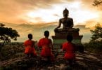 Três pequenos monges observando a estátua de um monge budista ao pôr-do-sol.