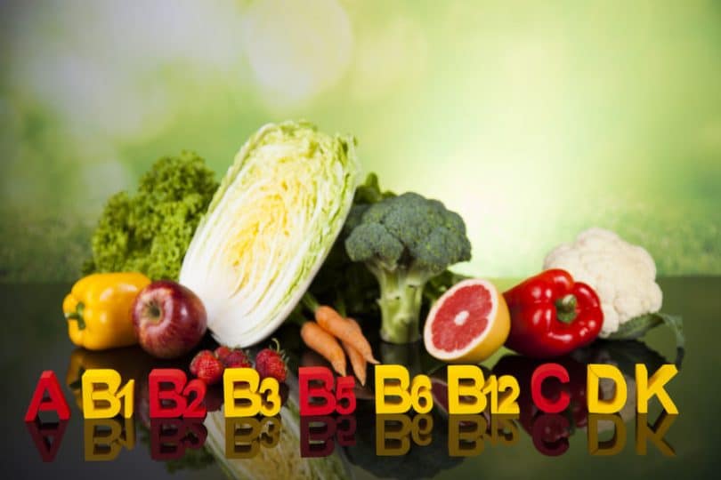 Alguns vegetais e frutas, com vitaminas escritas em fileira na frente deles. As vitaminas são: A, B1, B2, B3, B5, B6, B12, C, D e K.