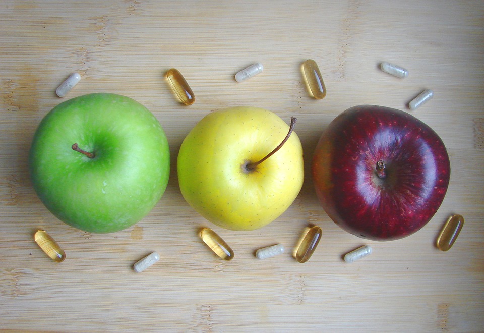 Três maçãs lado a lado, sendo uma verde, uma amarela e uma vermelha, vistas de cima. Ao redor delas estão distribuídas algumas pílulas.