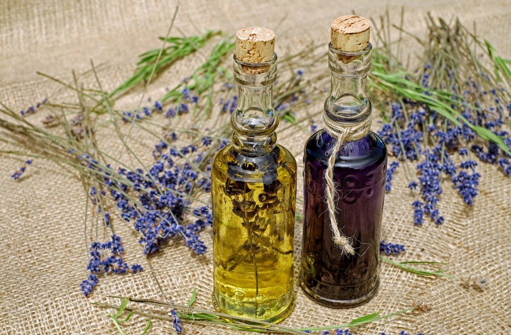 Duas garrafinhas com óleos essenciais, um roxo e um amarelo, fechadas com rolhas, Atrás das garrafas, ramos de uma planta roxa.