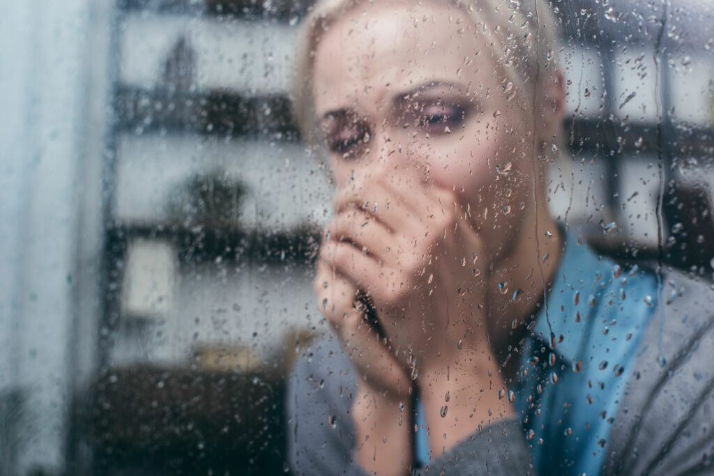 Mulher chorando com as mãos na frente do rosto, de frente para uma janela molhada de chuva.