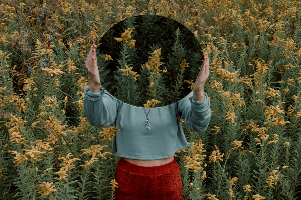 Mulher em um campo de flores segurando um espelho na frente da cabeça, a qual se torna invisível.