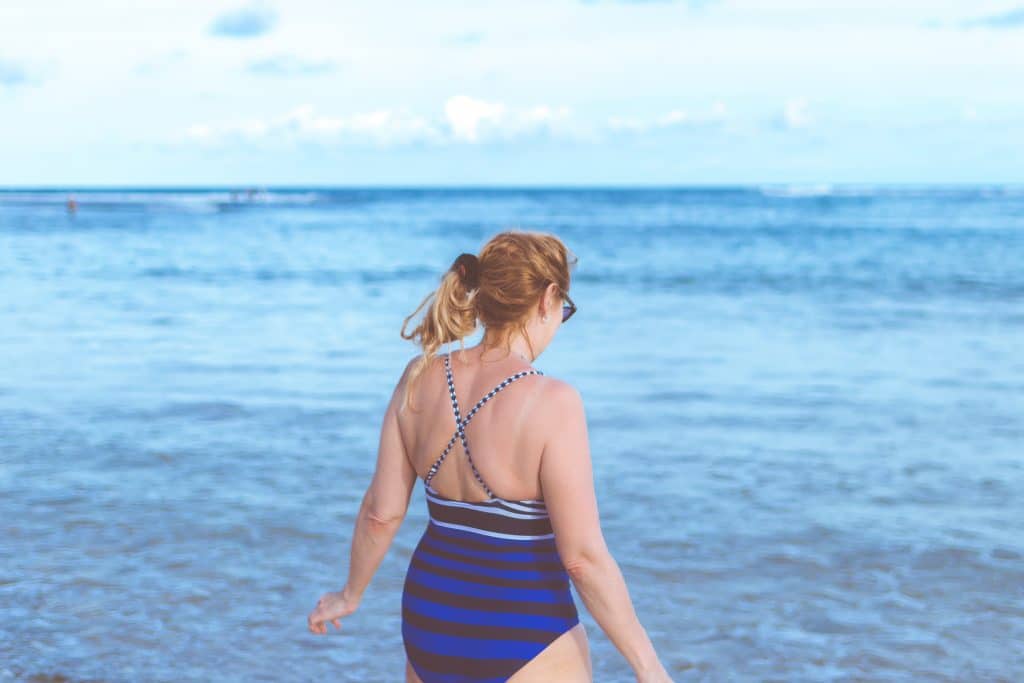 Mulher adulta usando maiô e com os cabelos presos de frente para a praia.