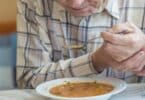 Imagem de um senhor de idade na frente de um prato de sopa levando uma colher para a boca com as duas mãos
