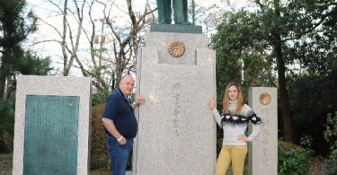 Um homem e uma mulher ao lado de uma estátua de um homem, com pedestal alto.