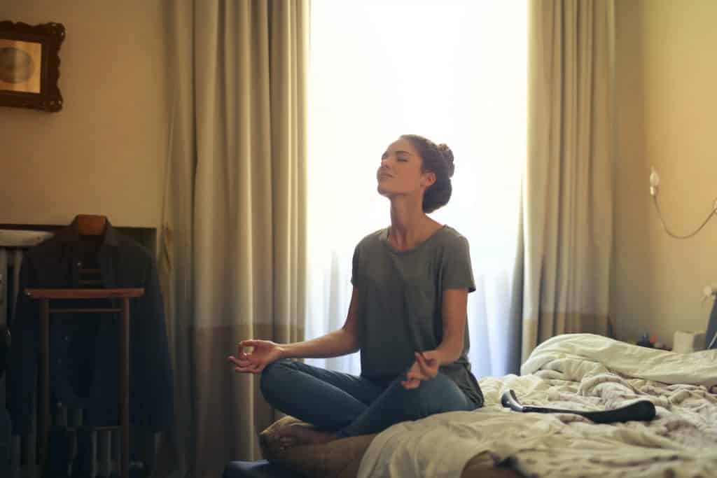 Mulher sentada em uma cama, meditando de olhos fechados.