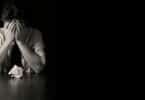 Imagem de um homem com as mãos no rosto em um cenário escuro, apoiado na mesa, como se estivesse tendo pensamentos ruins