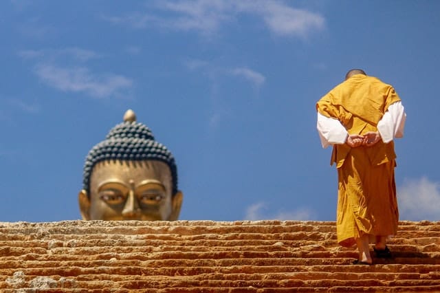 Monge subindo escadaria visto de costas com rosto de estátua de buda ao fundo