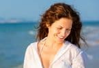 Mulher na praia sorrindo olhando para baixo com vento no cabelos