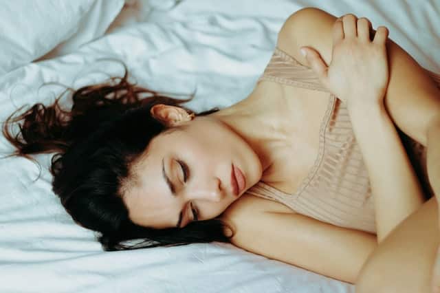 Mulher branca de cabelos castanhos deitada numa cama branca, com os braços ao entorno do corpo.