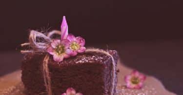 Imagem de um pedaço de brownie de chocolate disposto sobre uma travessa. Ele está decorado com um laço e um flor cor de rosa.