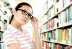 Mulher em uma biblioteca com olhar reflexivo e com uma mão ajustando o óculos