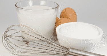 Leite em um copo de vidro, ovos, açúcar e um batedor manual de ovos sobre uma mesa