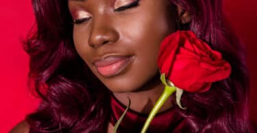 Mulher maquiada em tons de vermelho segurando uma rosa vermelha de olhos fechados