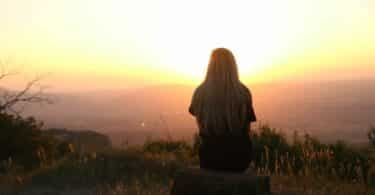 Imagem de uma mulher sentada de costas em uma rocha ao pôr-do-sol