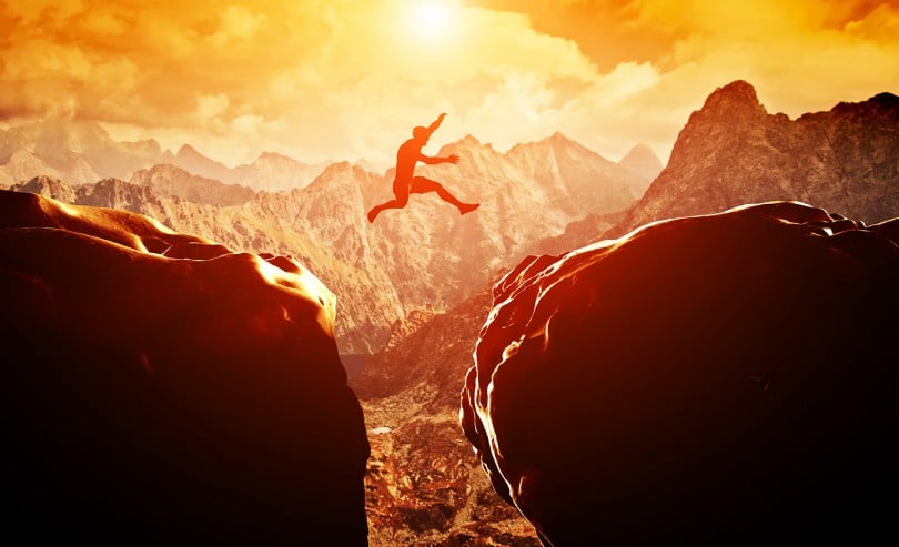 Homem pulando precipício entre duas montanhas rochosas ao pôr do sol. Liberdade, risco, desafio, sucesso.