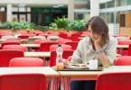 Mulher triste sentada em refeitório vazio, em frente a uma bandeja com comida intocada.