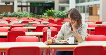 Mulher triste sentada em refeitório vazio, em frente a uma bandeja com comida intocada.