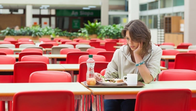 Mulher triste sentada em refeitório vazio, em frente a uma bandeja com comida intocada.