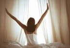 Mulher sentada em uma cama, de frente para uma janela com cortinas levemente abertas, e espreguiçando seus braços para cima.