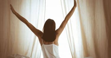 Mulher sentada em uma cama, de frente para uma janela com cortinas levemente abertas, e espreguiçando seus braços para cima.