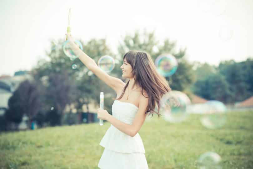 Mulher jovem sorrindo e brincando com bolhas de sabaão em um parque.