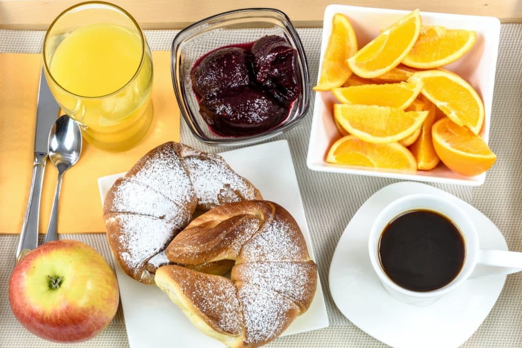 Mesa com pedaços de laranja, uma maçã, uma xícara de café, um copo de suco, e uma tigela com açaí.