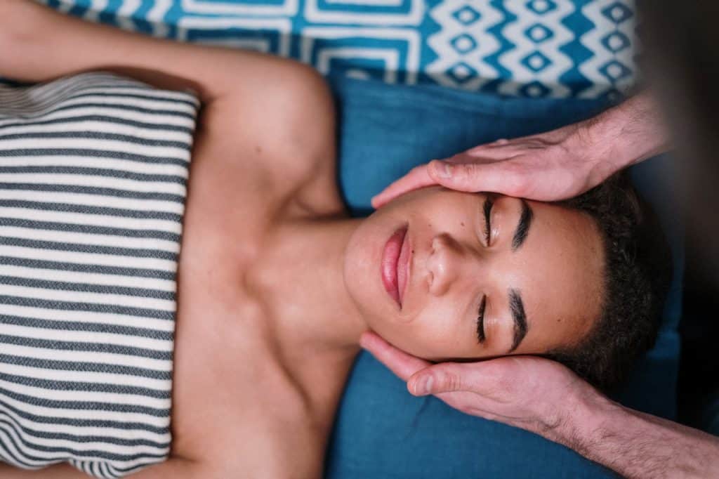 Mulher deitada com os olhos fechados enquanto recebe massagem facial. 