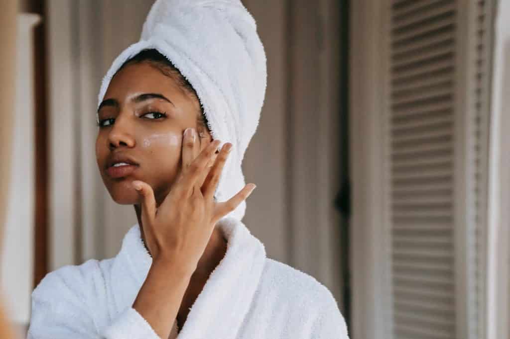 Mulher negra de roupão e toalha branca na cabeça fazendo massagem no rosto.