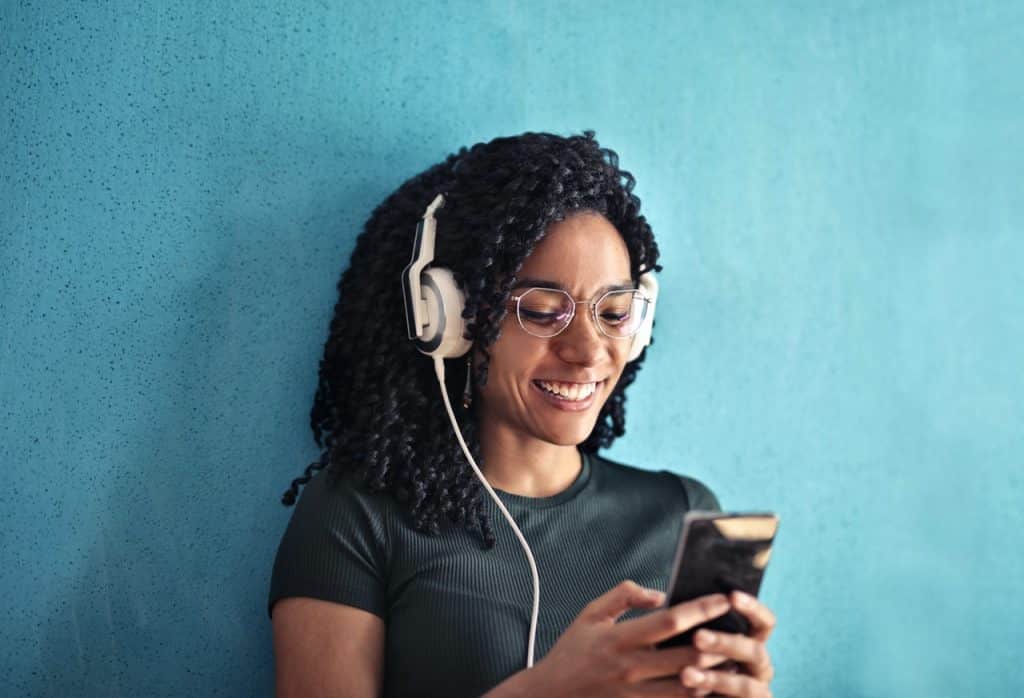 Mulher com fones do ouvindo, sorrindo, e digitando em um celular.