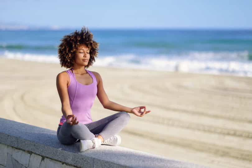 Mulher negra meditando com os olhos fechados na praia.