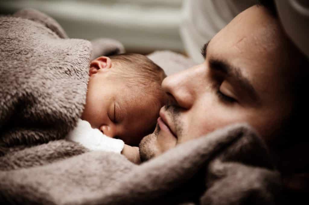 Pai deitado com um bebê no colo dormindo debaixo das cobertas. 