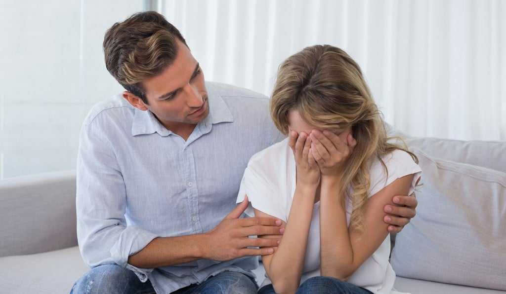 Homem consolando mulher que está chorando. Ele a envolve, com os braços ao redor dos ombros dela