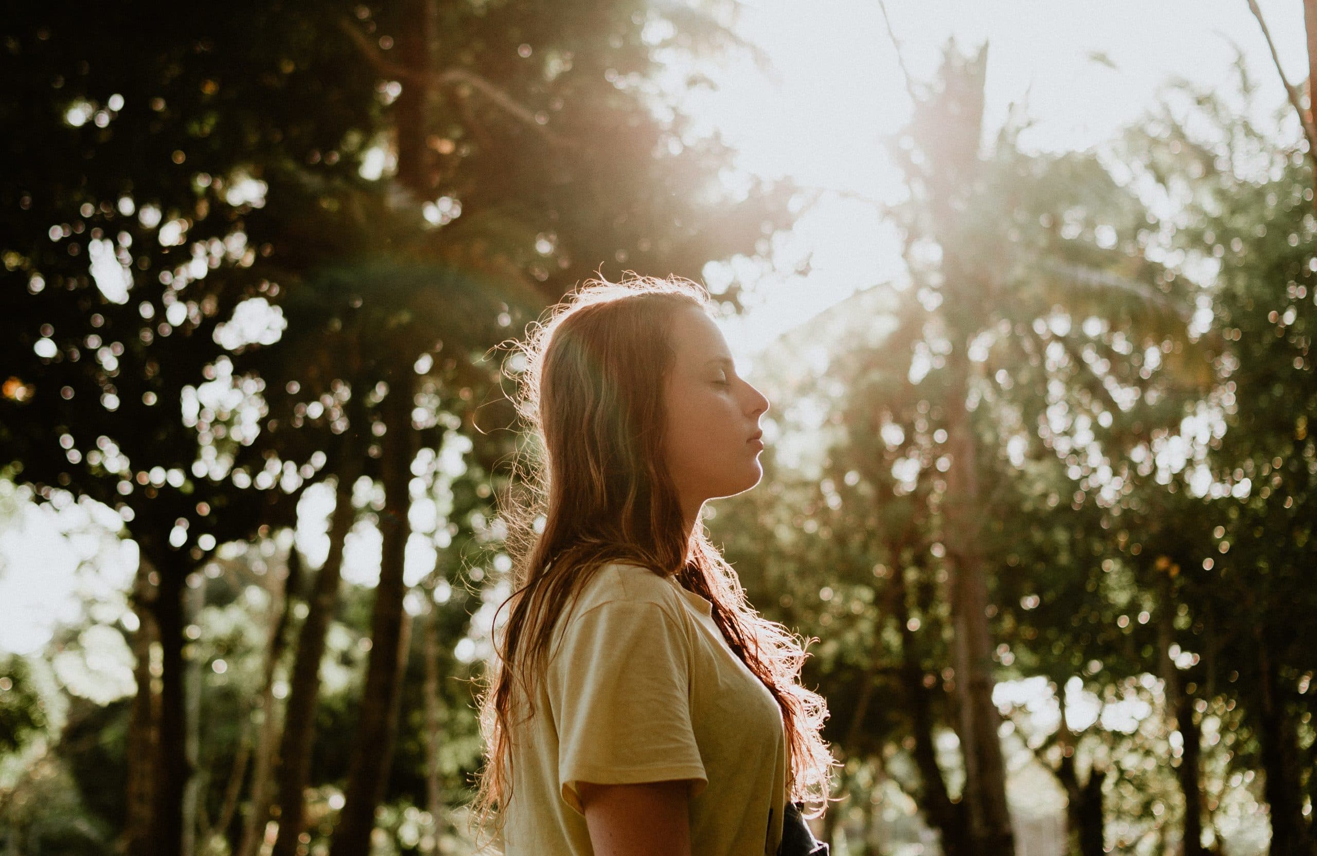 Mulher de olhos fechados com árvores e sol refletindo ao fundo