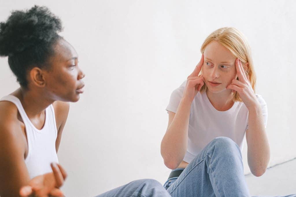 Duas mulheres sentadas conversando enquanto uma coloca os dedos sobre os lados de sua cabeça.