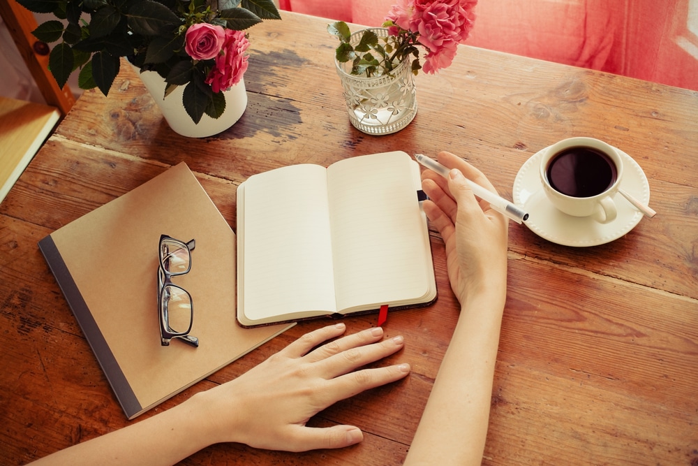 Pessoa escrevendo em caderno em uma mesa, com uma xícara de café e flores ao lado.