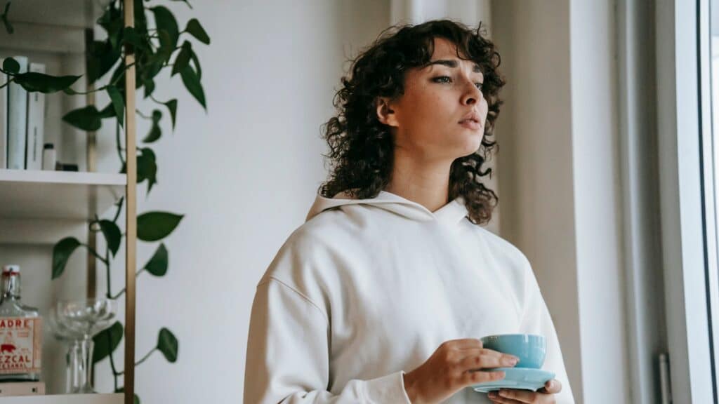 Mulher segurando xícara olhando pensativa para a janela