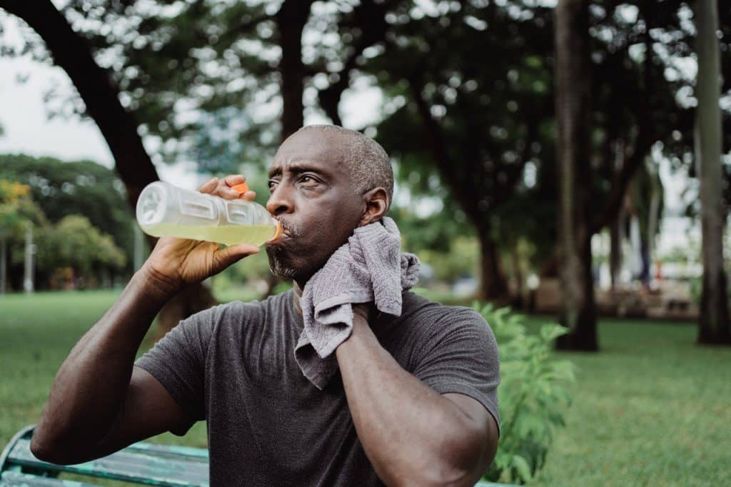 Homem negro tomando suco enquanto limpa o suor do rosto com uma toalha. 